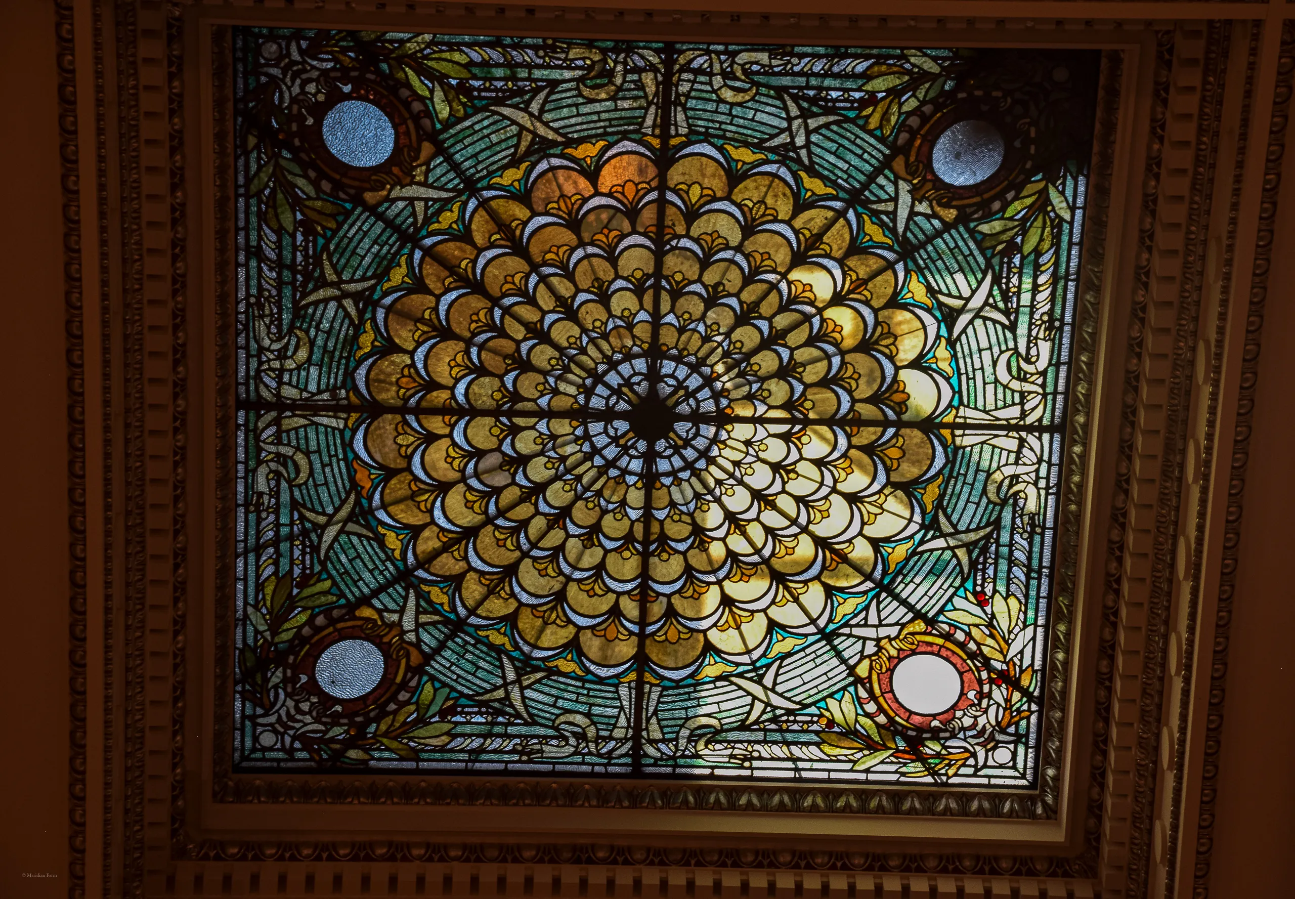 A square stained-glass skylight set in a carved coffered ceiling, its amber and blue-green glass arranged as a radial floral medallion with four small circular lights at the corners.